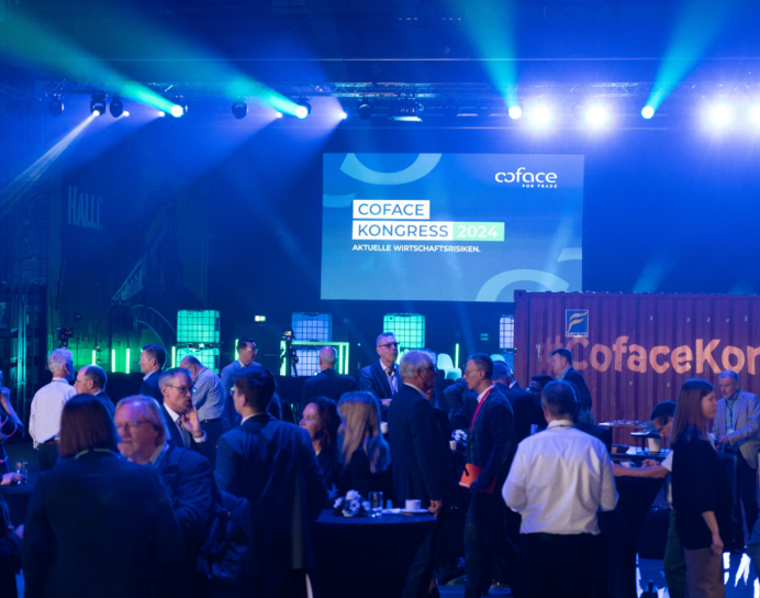 Dieses Bild zeigt einige Teilnehmende des Kongress im Vordergrund. Weiter hinten lässt sich die ausgeleuchtete Bühne mit einem dahinterliegenden Screen erkennen. Auf diesem Leinwand ist einen Folie eingeblendet. Auf dieser steht in Großbuchstaben "Coface Kongress 2024 - Aktuelle Wirtschaftsrisiken"