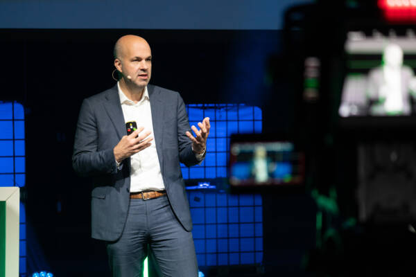 Dieses Bild zeigt Prof. Marcel Fratzscher, wie er während seiner Keynote auf der Bühne steht und zum anwesenden Publikum spricht. In seiner rechten Hand hält er das Steuerungselement für seine Präsentation. Er trägt einen dunkelblauen Anzug und ein weißes Businesshemd darunter.
